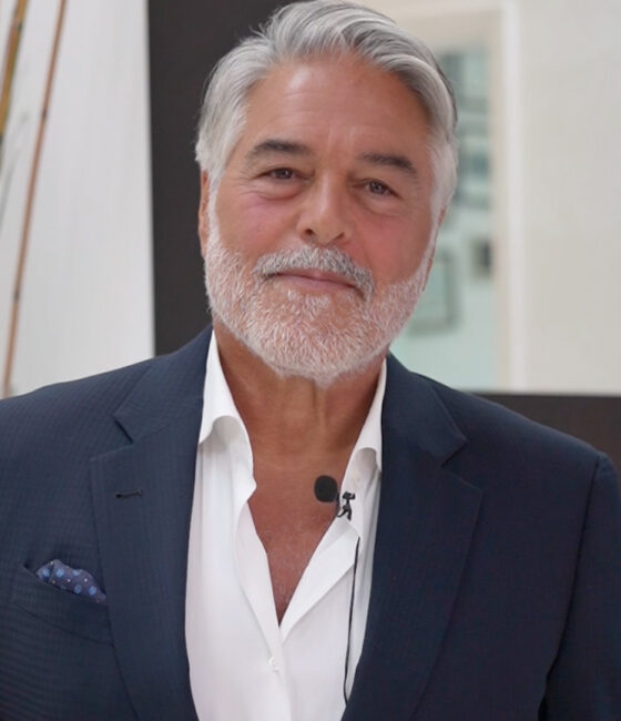 Smiling older man with gray hair and beard in a navy suit, white shirt, and a lavalier mic ready to speak.