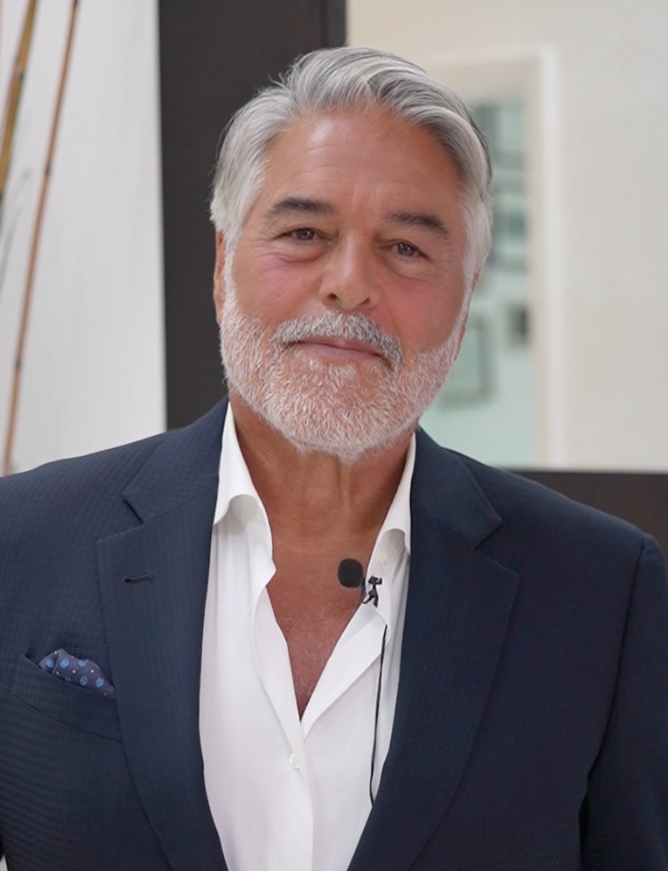 Chuck Alfieri Hair Consultation - Chuck Alfieri Smiling older man with gray hair and beard in a navy suit, white shirt, and a lavalier mic ready to speak.